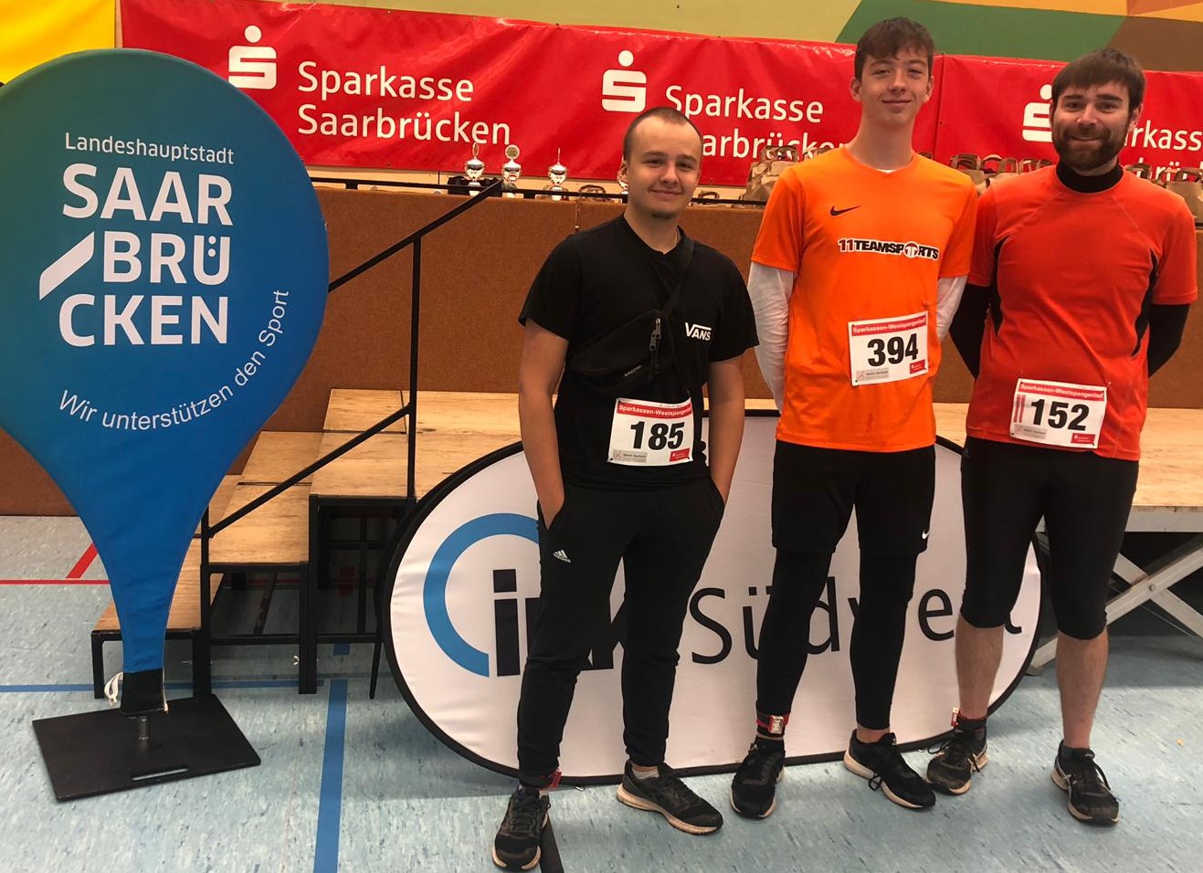 Drei Läufer mit Startnummern posieren beim Westspangenlauf vor Sparkasse- und Saarbrücken-Bannern.