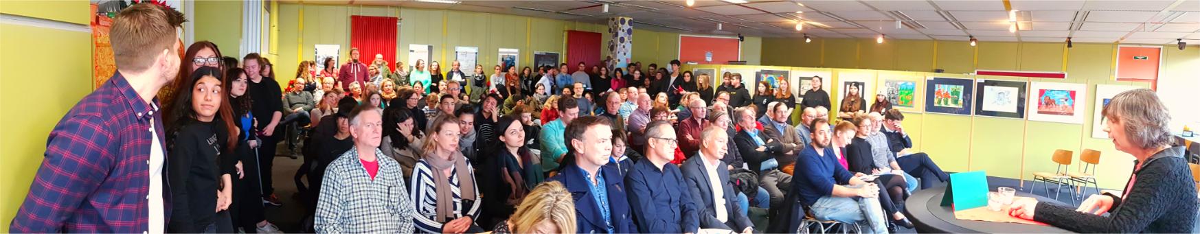 Publikum bei der Eröffnung der Kulturwoche im Schulcafé, an den Wänden Schülerkunstwerke.
