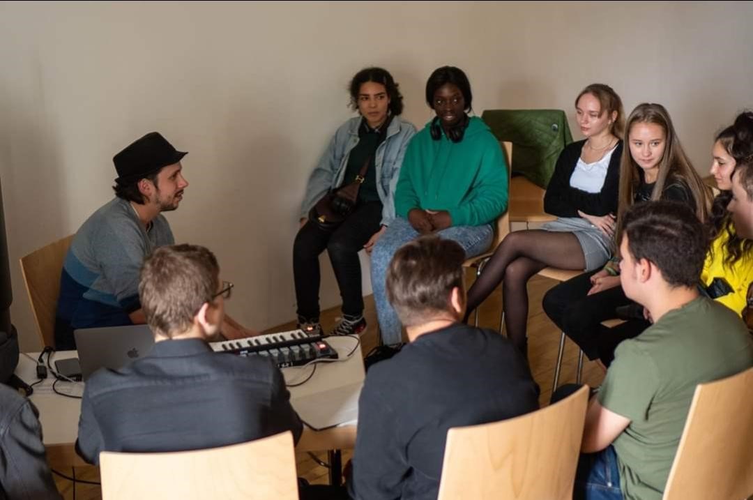 Ein Musiker mit Hut leitet am Keyboard einen Sitzkreis von Schülerinnen und Schülern.