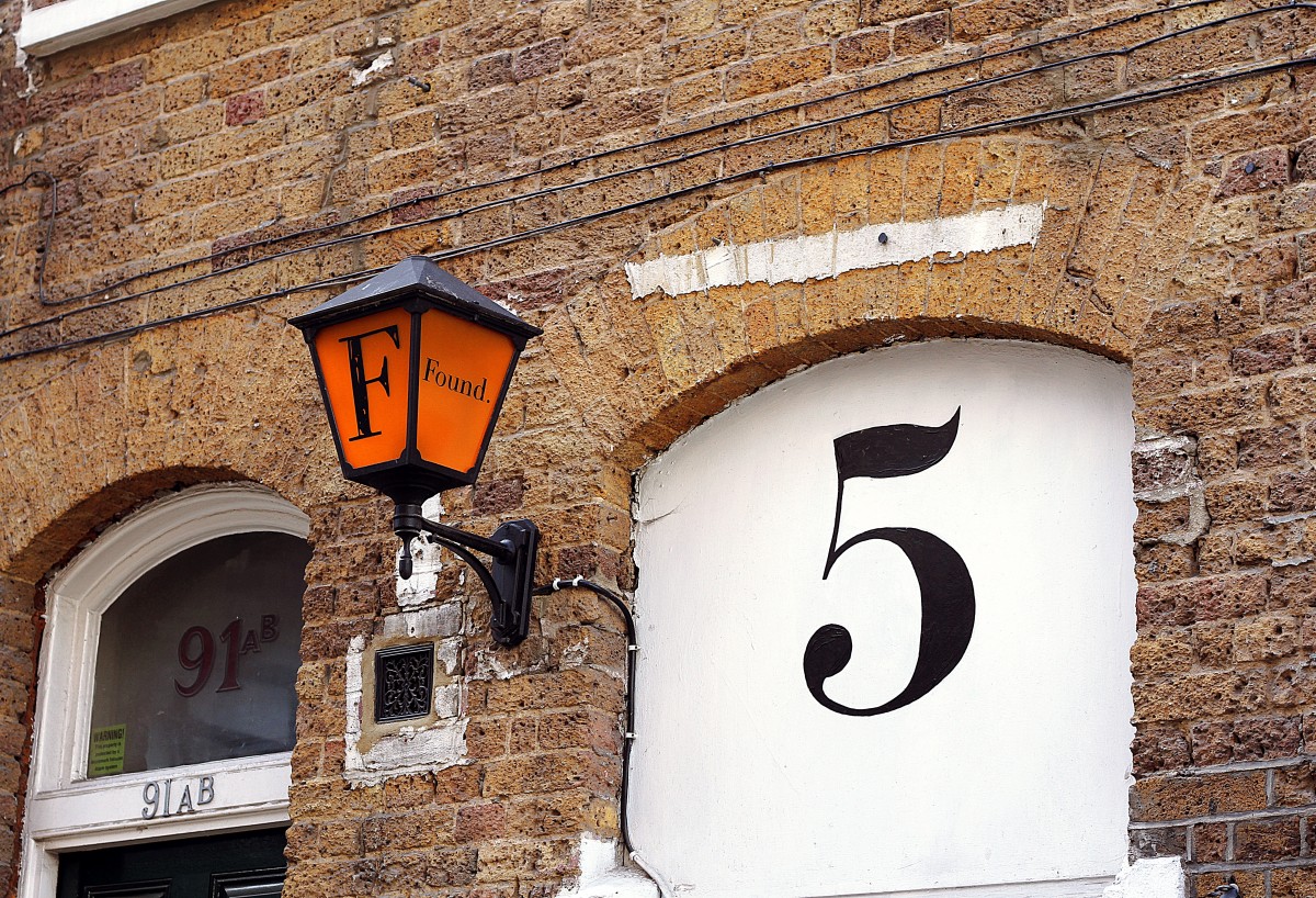 Große aufgemalte Hausnummer „5“ an einer Ziegelwand neben einer orangefarbenen „Found“-Laterne.