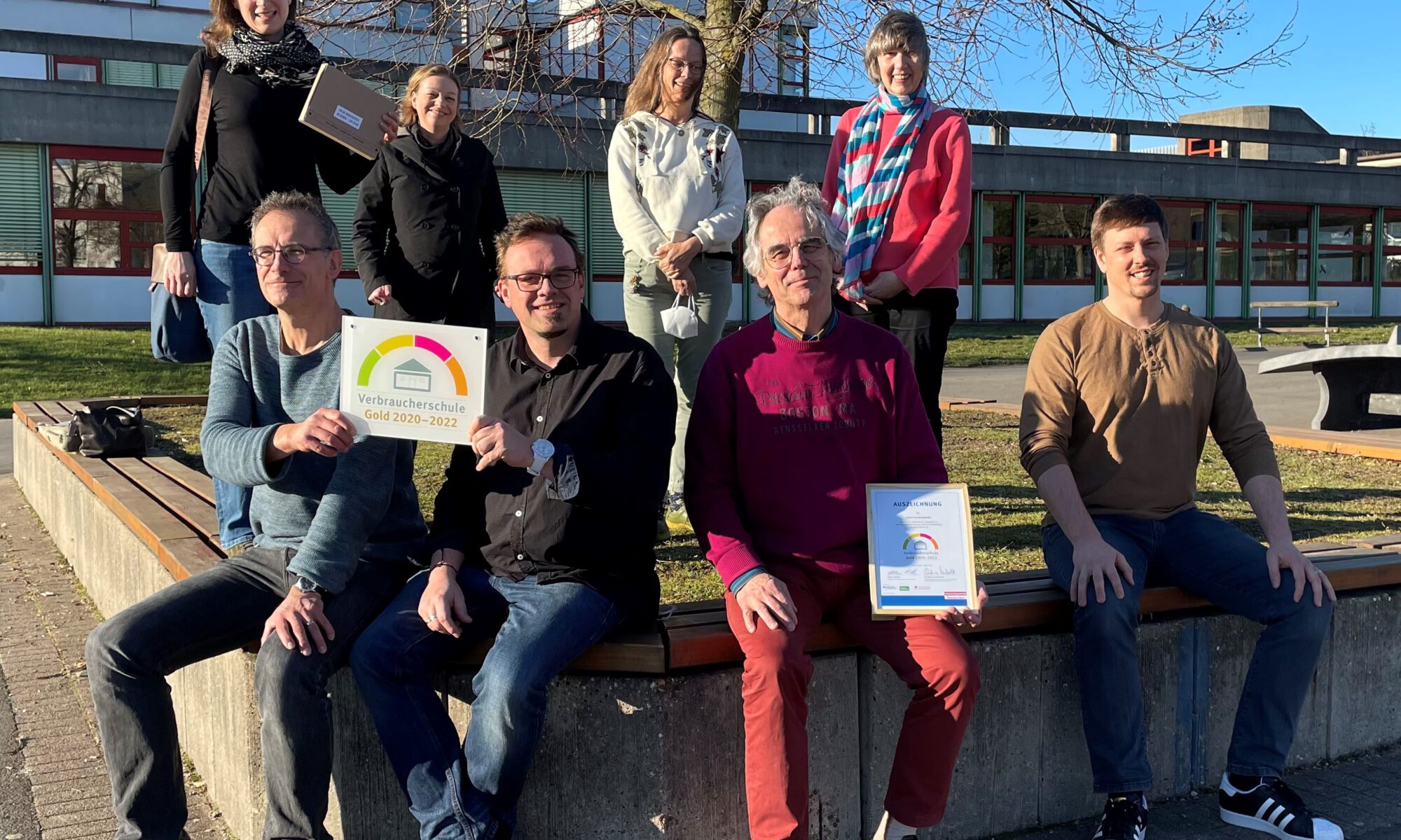 Acht Lehrkräfte sitzen im Freien auf einer Bank und halten das Schild „Verbraucherschule Gold 2020–2022“ und die Urkunde.