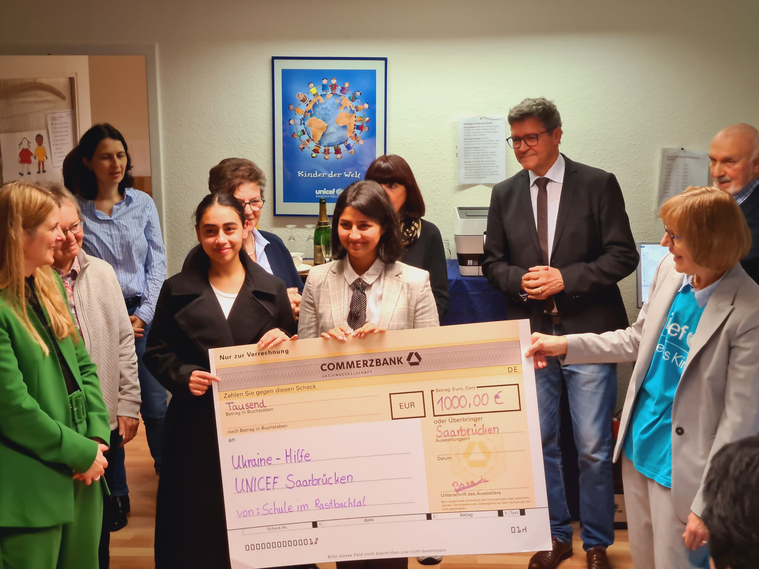 Zwei Schülerinnen überreichen einen Spendenscheck über 1000 Euro für die UNICEF-Ukraine-Hilfe, umringt von Erwachsenen; im Hintergrund ein UNICEF-Plakat.