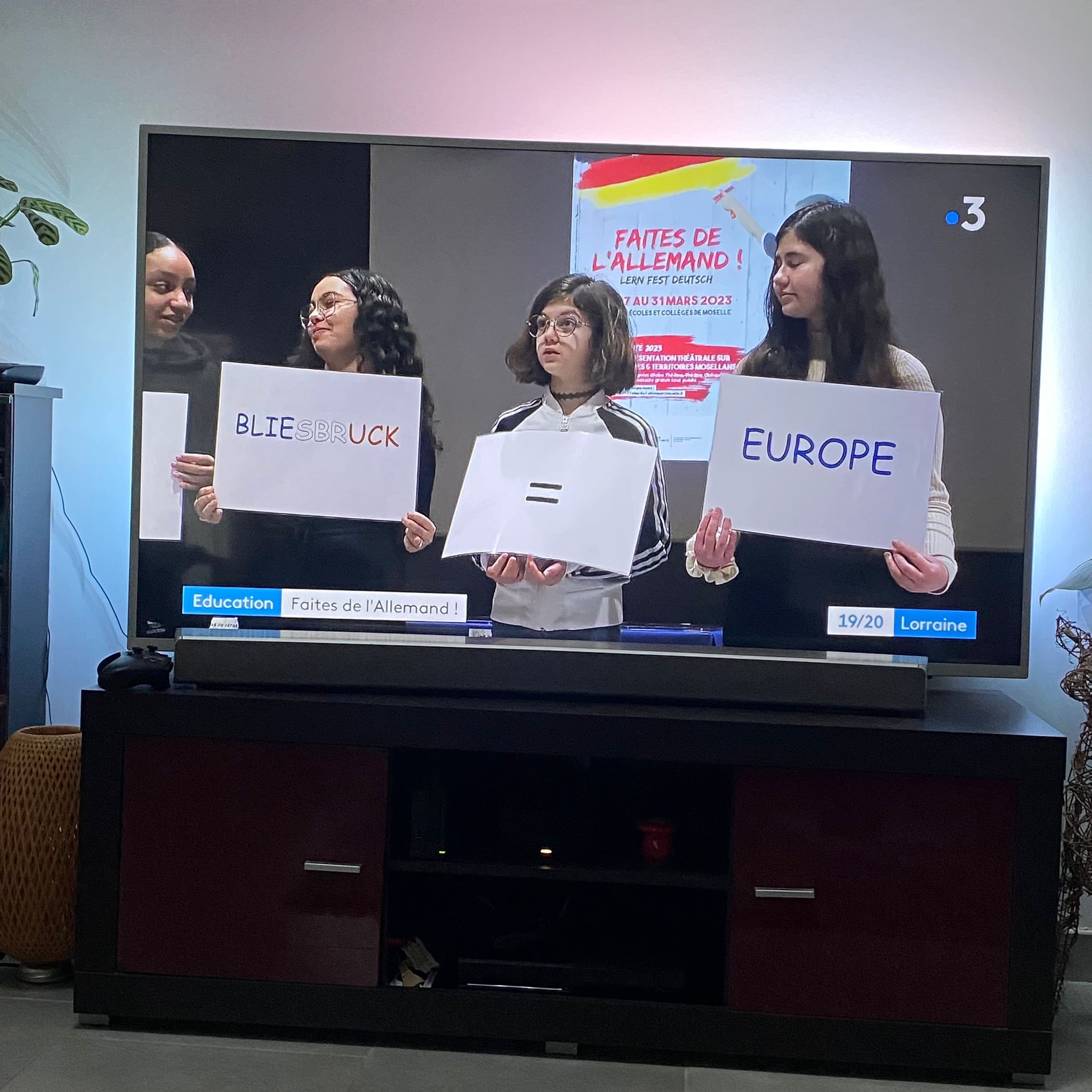 Ein Fernseher zeigt einen France-3-Beitrag „Faites de l’Allemand!“; Schülerinnen halten Schilder „Bliesbruck = Europe“.