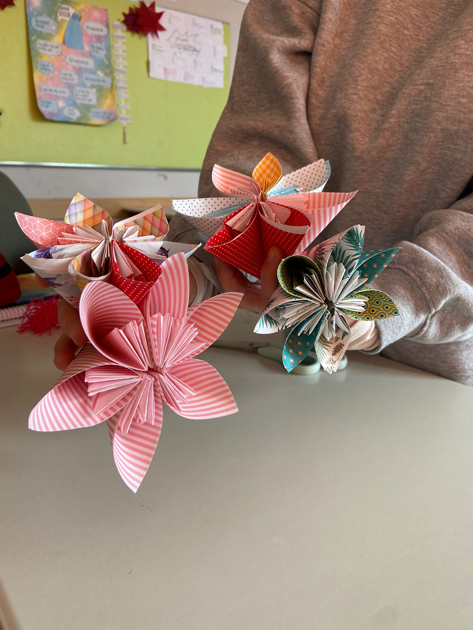 Zwei Hände halten selbst gefaltete Papierblumen in Rosa und Rot im Klassenraum.