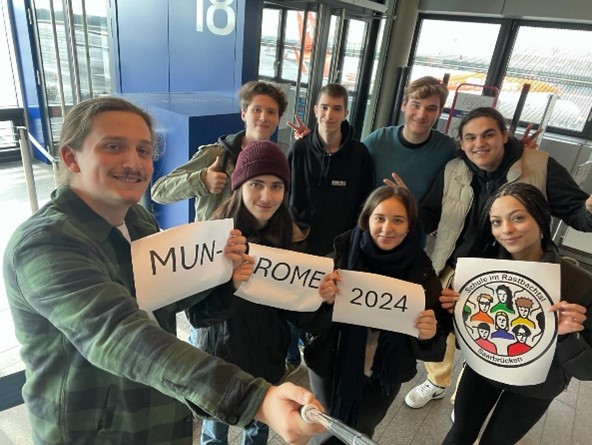 Selfie der Schülergruppe am Flughafen mit selbst gemalten Schildern „MUN ROME 2024“ und dem Schullogo.