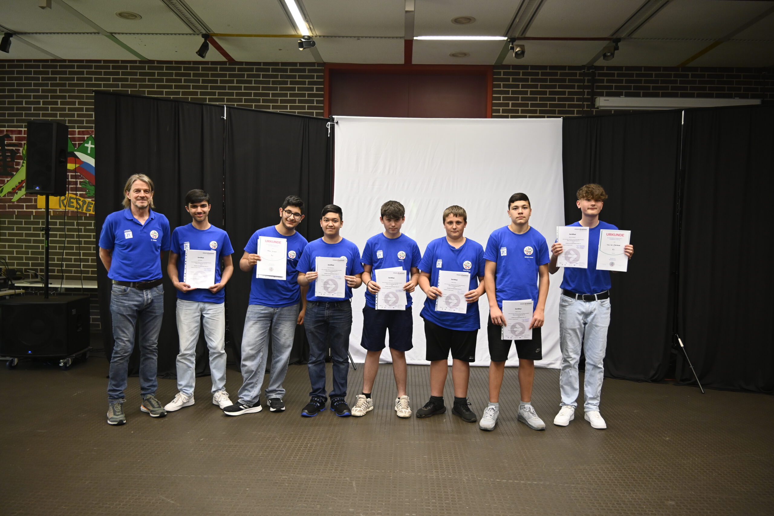 Sieben Schüler in blauen Shirts halten mit einem Lehrer ihre Urkunden auf der Bühne.