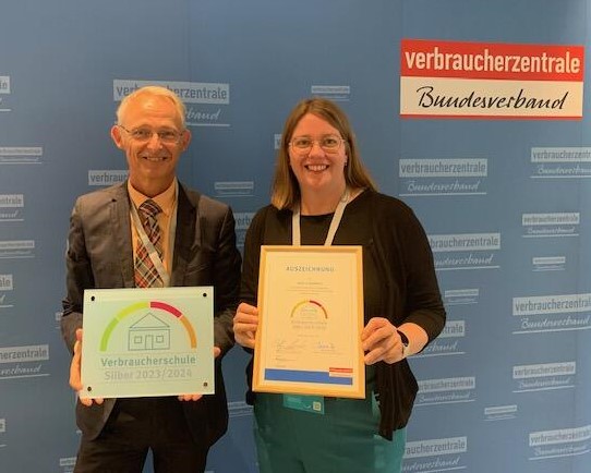 Ein Mann und eine Frau halten vor einer Verbraucherzentrale-Wand die Plakette „Verbraucherschule Silber 2023/2024“ und die gerahmte Urkunde.