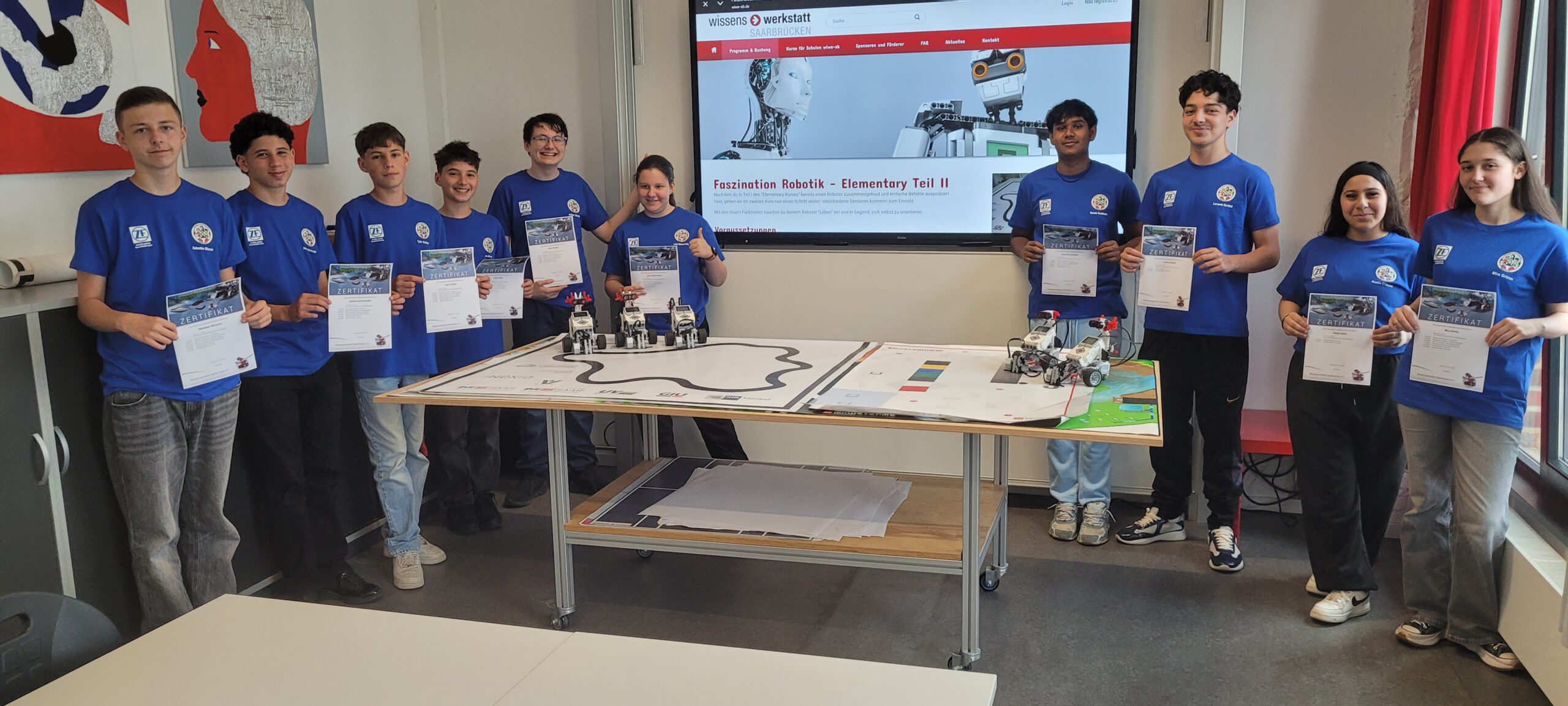Schülerinnen und Schüler in blauen ZF-T-Shirts halten ihre Praktikumszertifikate hoch; im Hintergrund ein Bildschirm zum Projekt „Faszination Robotik“.