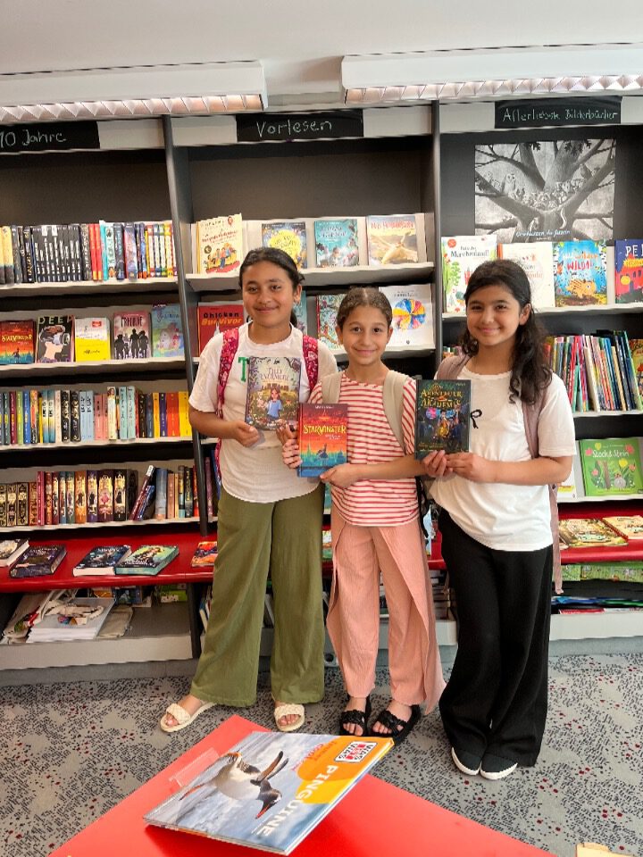 Drei Schülerinnen der Lese-AG halten in einer Buchhandlung ihre selbst ausgesuchten Bücher in die Kamera; im Hintergrund Regale mit Kinderbüchern.